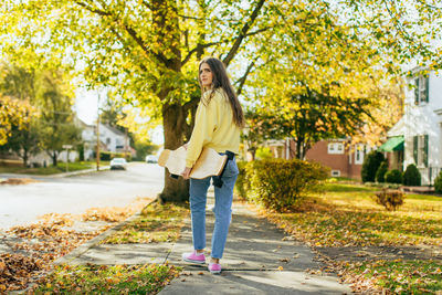 Girl with long hair holding longboard while walking on sidewalk in neighborhood looking at camera 