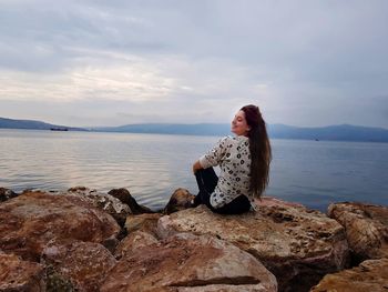 Rear view of smiling young woman sitting on rock against cloudy sky during sunset