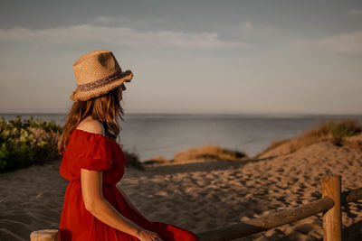 Side view of woman wearing hat sitting against sea and sky