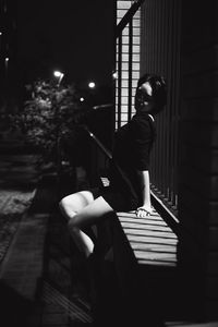 High angle view of young woman sitting on wall with fence at night