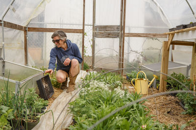 A smiling woman in cowboy boots tends to seedlings in a greenhouse