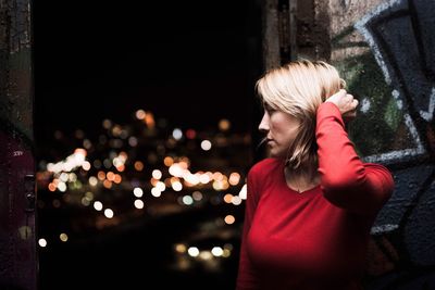 Thoughtful woman standing by window against illuminated buildings