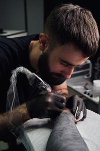 Concentrated male tattooist in gloves making tattoo on hand of client while using professional tattoo machine in modern tattoo studio