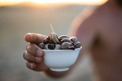 Midsection of shirtless man holding seashells in bowl at beach during sunset