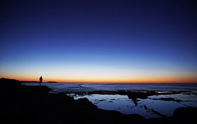 Silhouette person standing on shore against clear blue sky during sunset