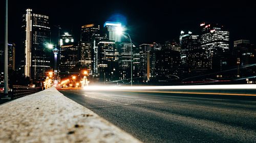 Illuminated light trails on road by buildings against sky at night