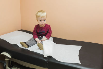 High angle view of baby boy with paper looking away while sitting on bed at hospital
