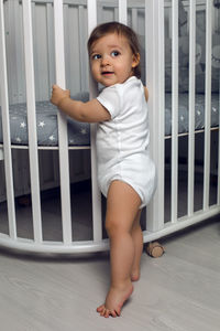 Child in white clothes of one year stands on the floor next to his round white bed