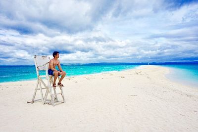 Shirtless young man sitting on lifeguard chair at beach against cloudy sky