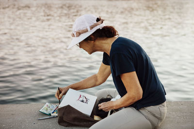 Woman painting while sitting on retaining wall by sea