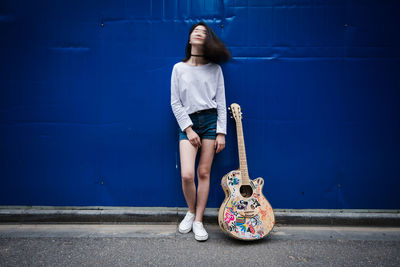 Full length of young woman standing by guitar against blue wall