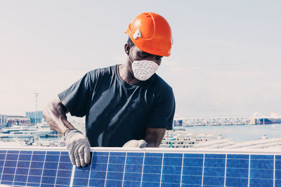 Black workman in helmet and protective mask installing solar battery in industrial area of contemporary plant