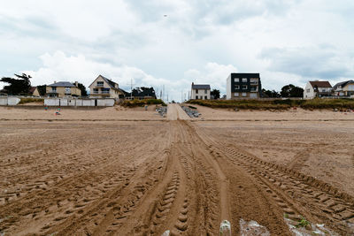 Tire tracks on dirt road by buildings against sky