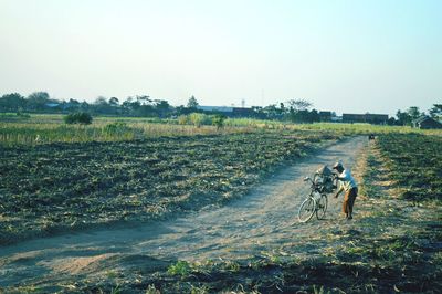 Young man with bicycle on dirt road against clear sky