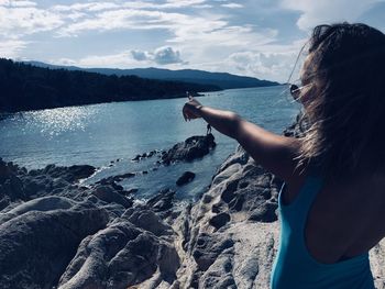 Young woman pointing at mountains by sea against sky during sunny day