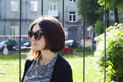 Young woman wearing sunglasses while standing by fence