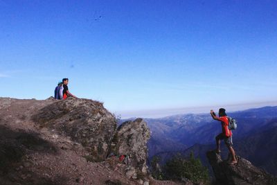 Full length of hiker photographing friends sitting on mountain against clear blue sky