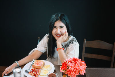 Portrait of smiling woman with breakfast on table sitting against black background