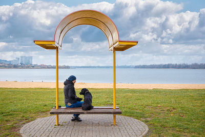 A girl with a black dog labrodor is sitting on a bench by the lake.
