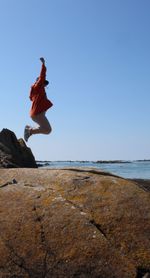 Side view of woman jumping on rock at beach against clear sky