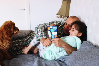 Man with daughter by dog reading book on bed at home