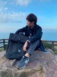 Young man sitting on bench against sky