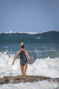 Rear view of woman with surfboard standing on rock at sea
