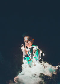 Young man with illuminated string lights and smoke standing against sky at night