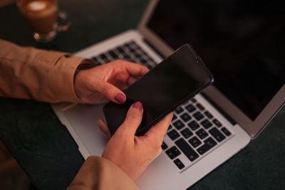 High angle of cropped unrecognizable young female freelancer messaging on mobile phone while sitting at table with laptop and cup of coffee in cozy cafe and working or remote project