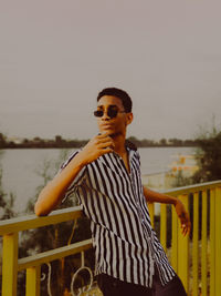 Man wearing sunglasses standing by railing against sky