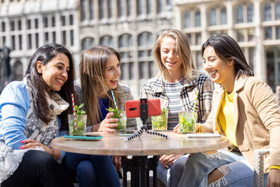 Four multi ethnic female friends or students at a cafe in the city using smartphone for a video call