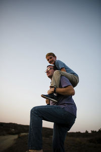 Low angle view of father carrying son on shoulders against clear sky during sunset