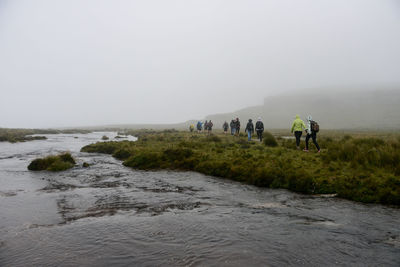 Hikers walking on field by lake during foggy weather