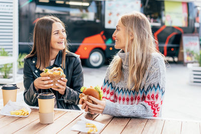Two middle-aged women eat fast food and chat at a street food market.