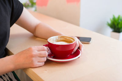 Red cup of coffee in hands on a table with mobile phone in cafe.