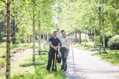 Full length portrait of smiling female trainee standing with male instructor holding gardening equipment on footpath at