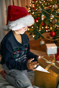 A happy boy in a santa claus hat opens a box with a christmas present next to a christmas tree 