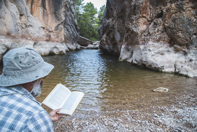 Tourist with a beard and hat reads a book sitting on the river bank