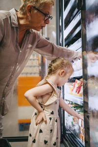 Side view of girl with hand on hip doing shopping from refrigerated section near grandmother at store