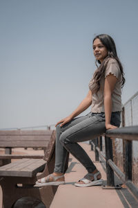 Portrait of young woman sitting on bench against clear sky