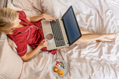 Directly above shot of woman holding coffee cup while using laptop on bed