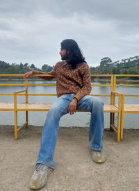 A long haired and bearded young man looking sideways while sitting on bench next to the lake