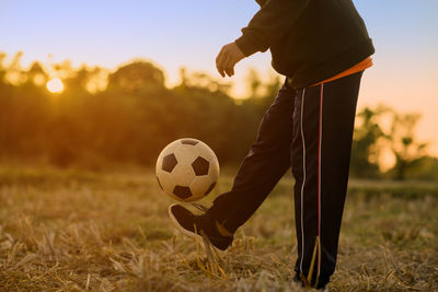 Low section of man playing soccer on field against sky during sunset