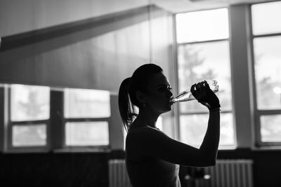 Side view of young woman drinking glass window at home