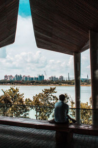 Rear view of woman sitting by sea against sky in city
