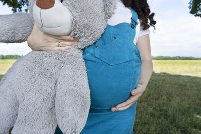 Young pregnant woman in blue sundress with a big teddy bear walking in the park.