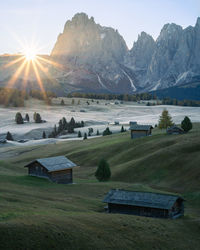 Vertical shot of sun rising above high alpine pasture with cottages from behind a wall of mountains