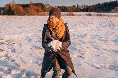 A woman in winter clothes collects a lump of snow in a snow-covered field