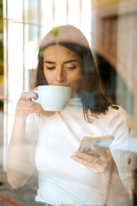 Through glass view of cheerful young female browsing mobile phone while standing near window with cup of coffee and enjoying free time at home