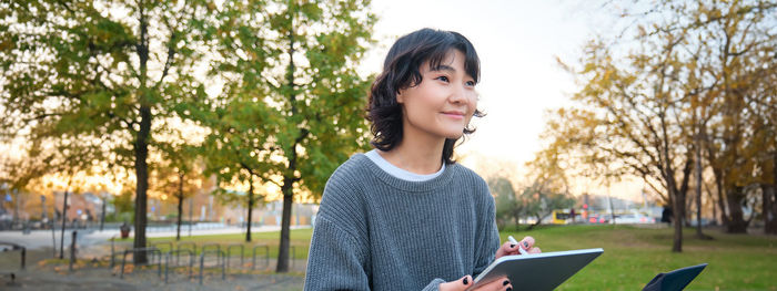 Portrait of young woman looking away while standing against trees